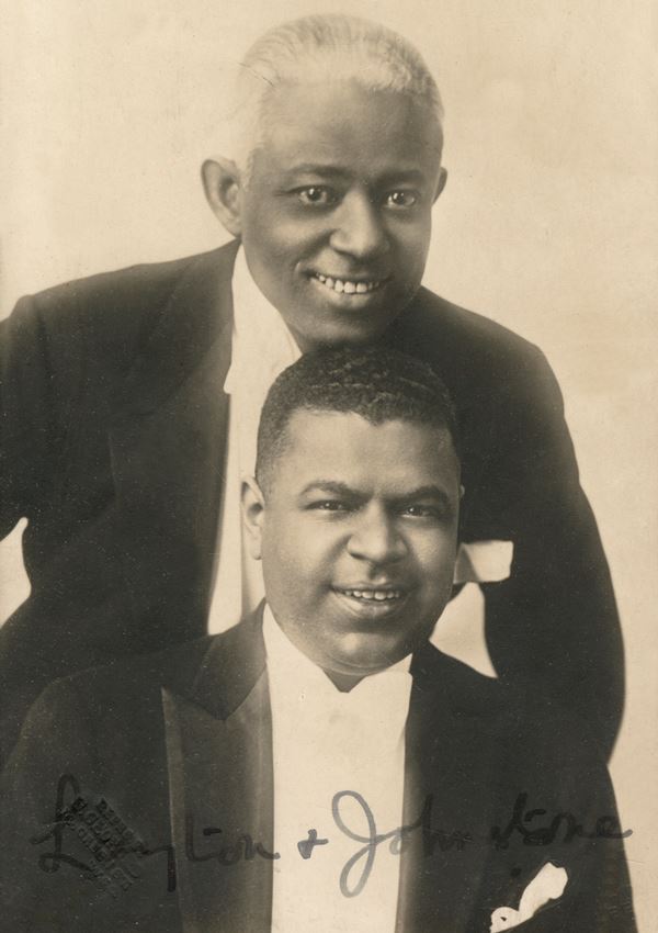 Black and white portrait photo of two black men posing together, the younger one sitting down and the older one standing behind him.